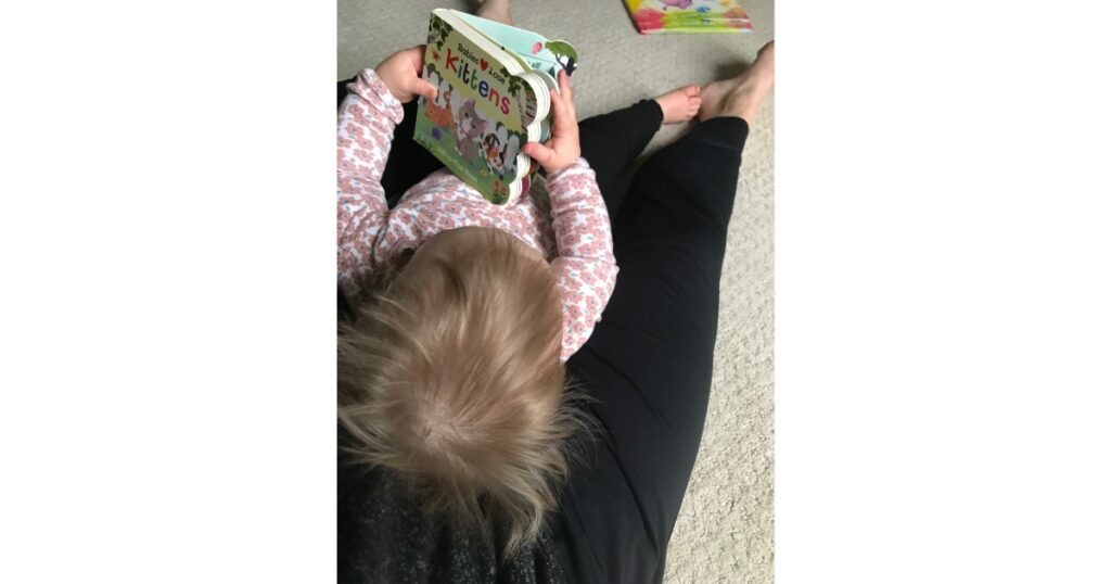 Child with blonde hair and pink flowered shirt laying on mom holding 'Babies Love Kittens' book.