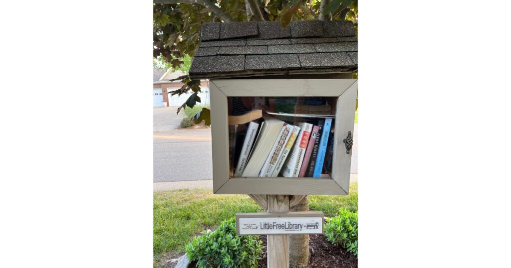 Little Free Library with books inside and placed in front of a tree and green grass beneath.