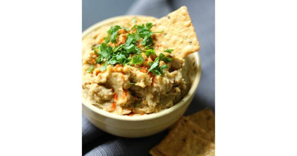 Yellow colored dip with cilantro on top and cracker in the bowl.