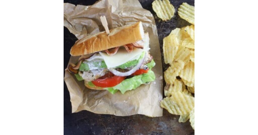 Sandiwch with multiple layers of tomato, onion, lettuce, chicken and bacon. Sandwich on parchment paper and a sandwich toothpick in it. Ruffle chips to the right.