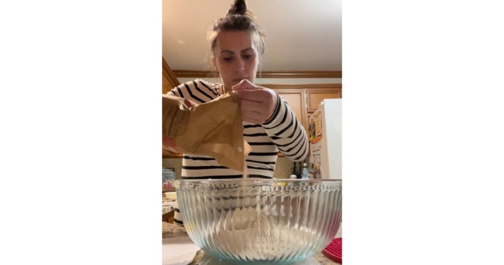 Woman in black and white stripped shirt pouring flour into a glass bowl on top of a scale.