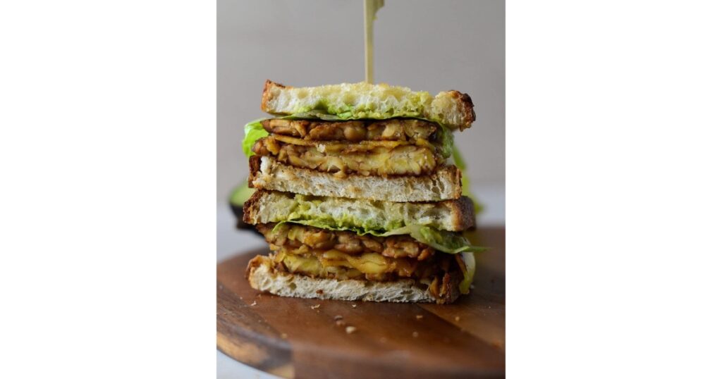 Stacked sandwich with tempeh and avocado. Sandwich toothpick on top. Sandwich atop a brown cutting board.