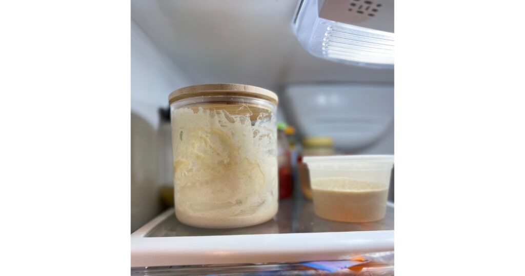 Sourdough in tall clear jar with a small container on right hand side in refrigerator