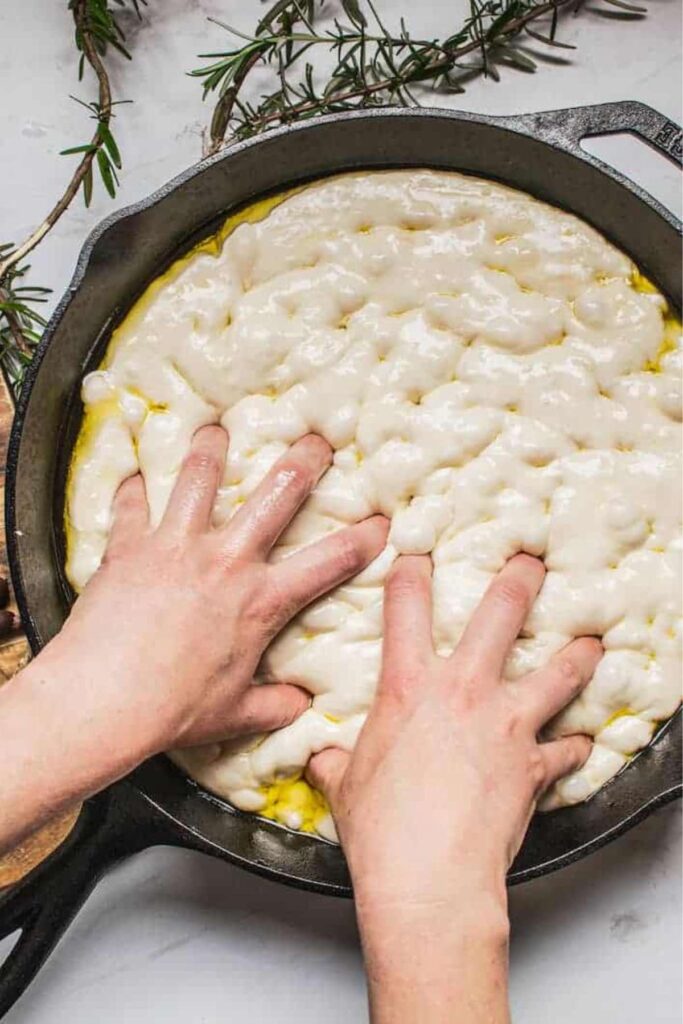 sourdough focaccia in cast iron skillet with hands making indents.