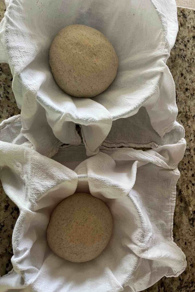 two sourdough boules in towels in bowls on counter.